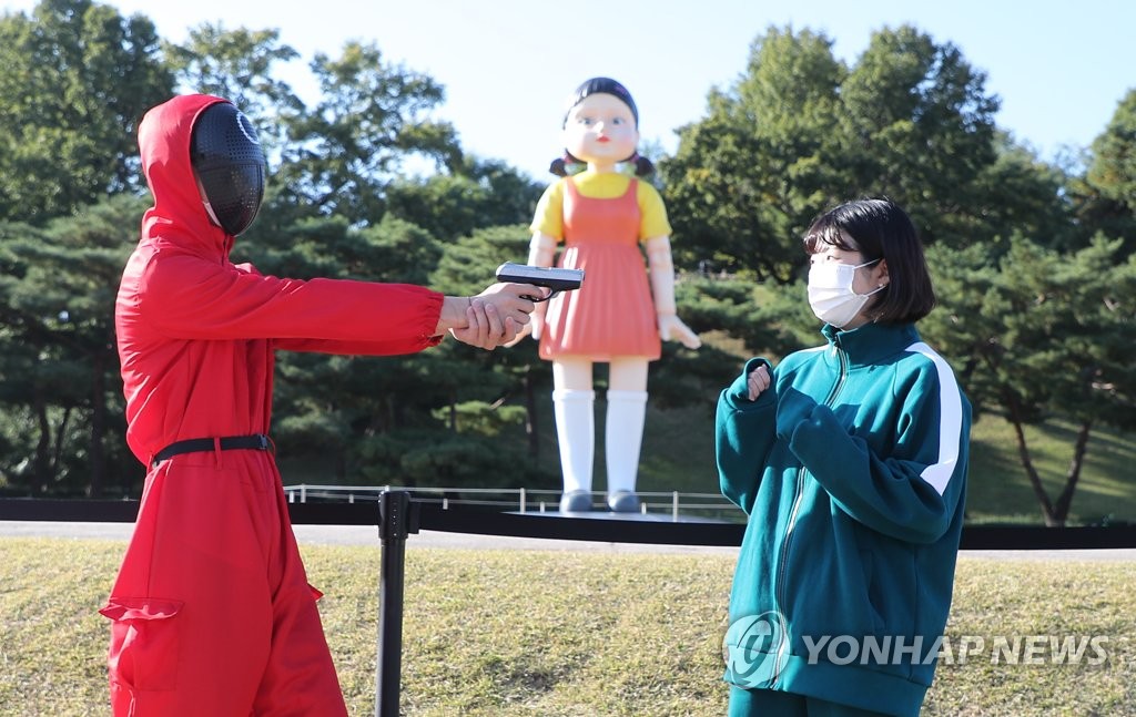 10月27日，在设于首尔松坡区奥林匹克公园的奈飞原创剧《鱿鱼游戏》木头人游戏机器人娃娃前，身穿同款剧中服装的市民打卡拍照。 韩联社